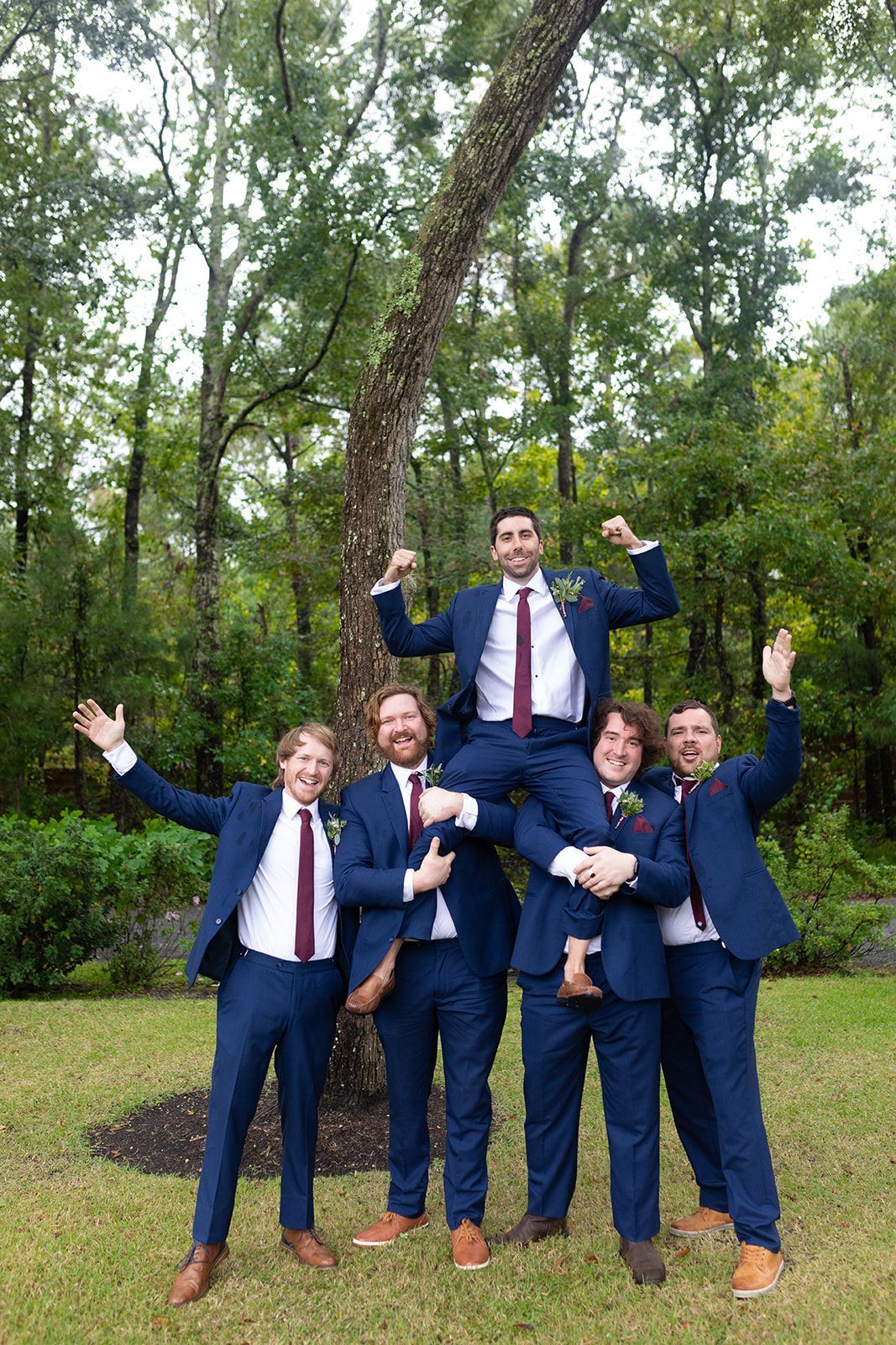 Five people in navy suits and red ties posing outdoors in a wooded area, smiling and cheering.