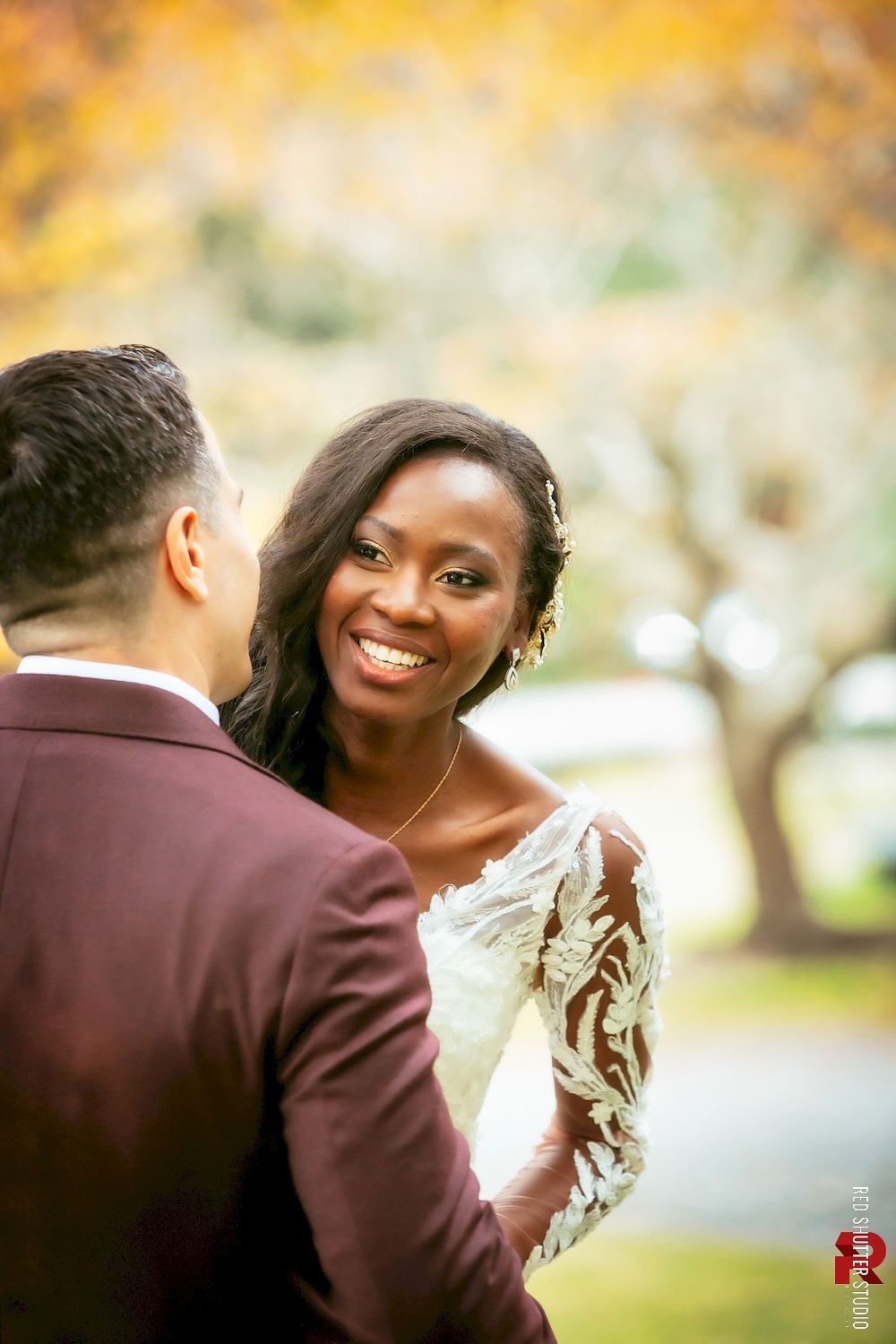 Smiling couple in wedding attire outdoors, with soft golden trees in the background