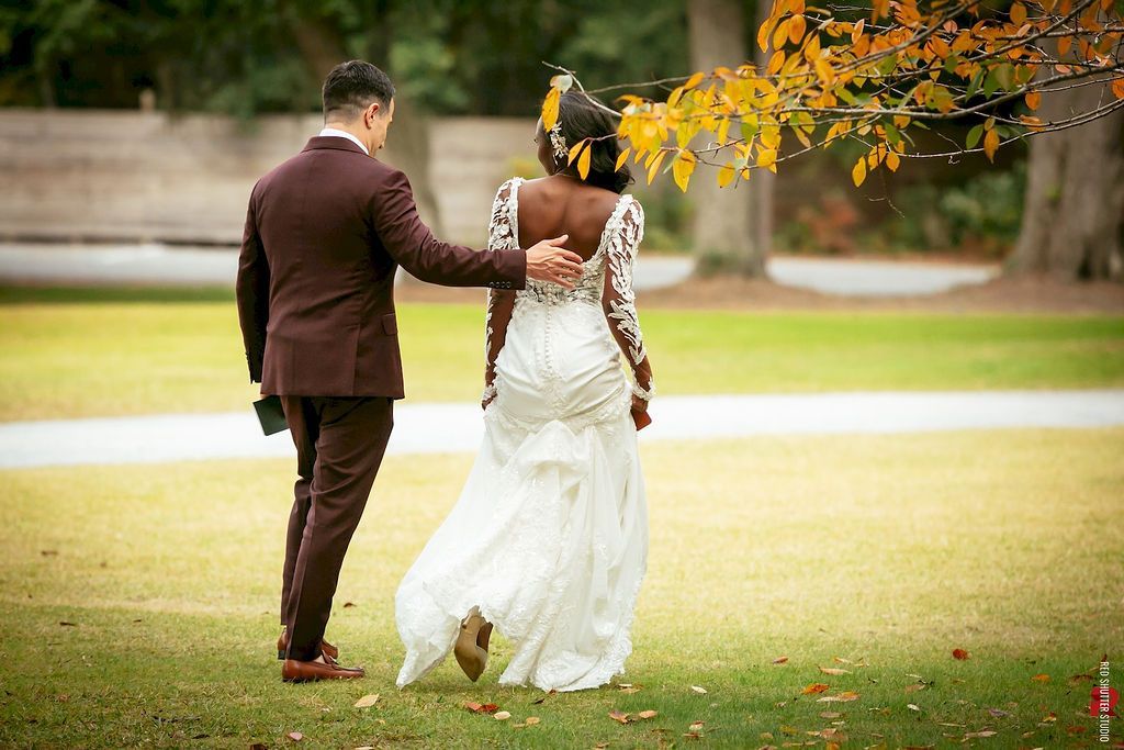 Couple walking hand in hand on a lawn, with autumn leaves overhead, the woman in a white dress and the man in a brown suit