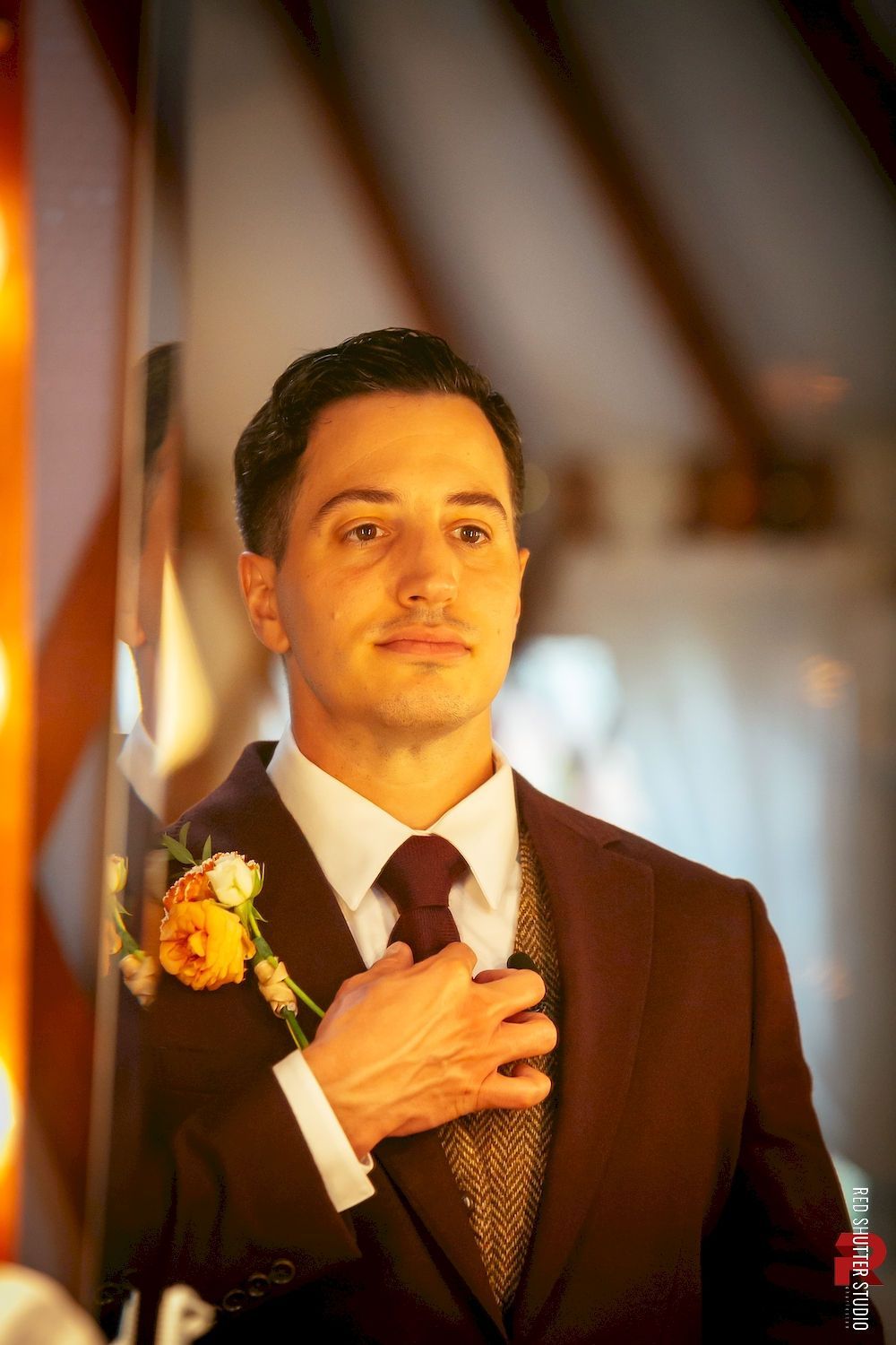 Man in a dark suit with a boutonniere, hand over heart, standing in warm indoor light