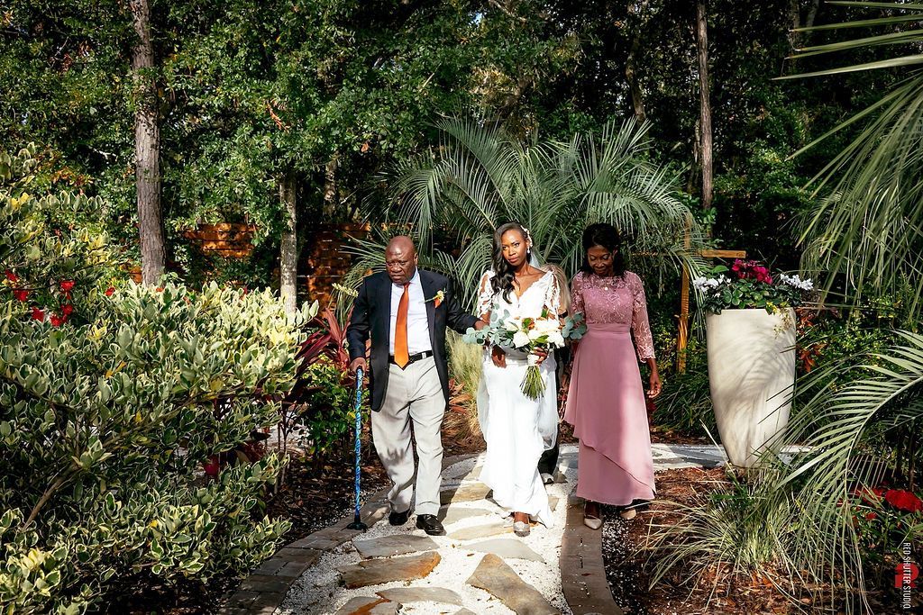 Three people walk along a garden path through lush greenery, one holding a bouquet.