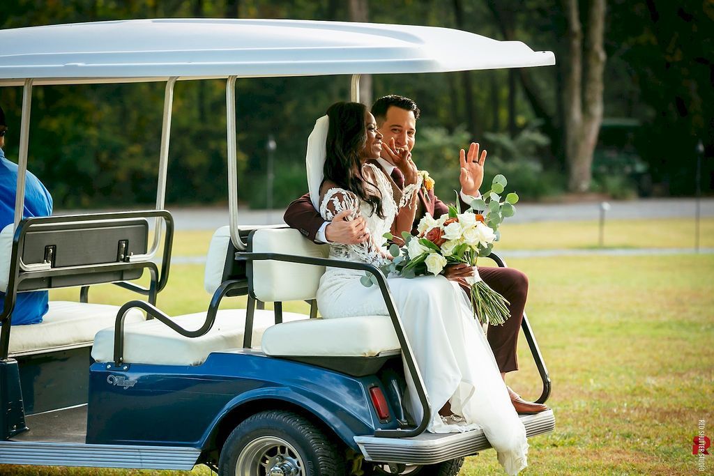Couple in white outfits riding a blue golf cart, smiling with a floral bouquet on a grassy lawn.