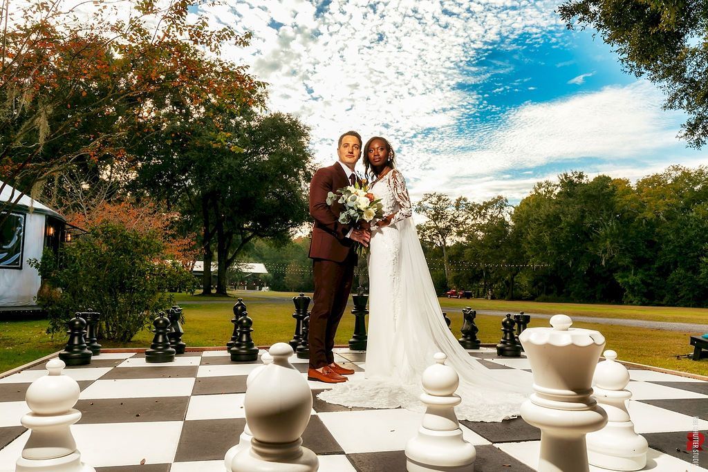 Couple in wedding attire standing on a giant outdoor chessboard in a garden.