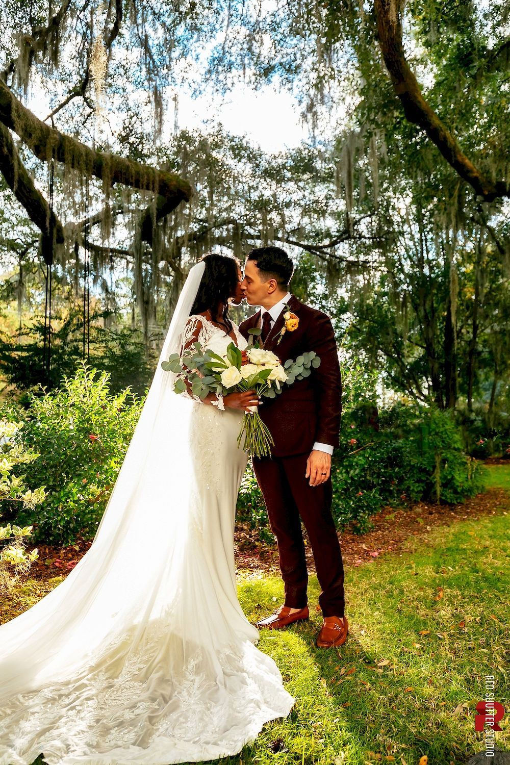 Bride and groom kissing in a garden, bride in white gown with long veil and bouquet, groom in dark suit