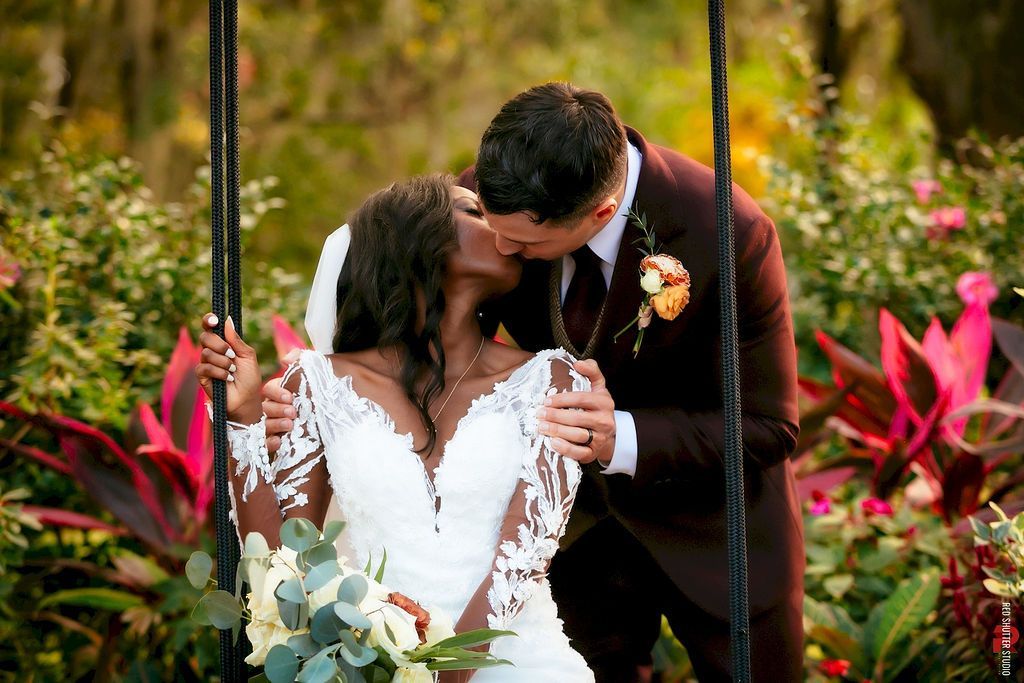 Couple kissing on a swing amid lush greenery and bright flowers at an outdoor wedding