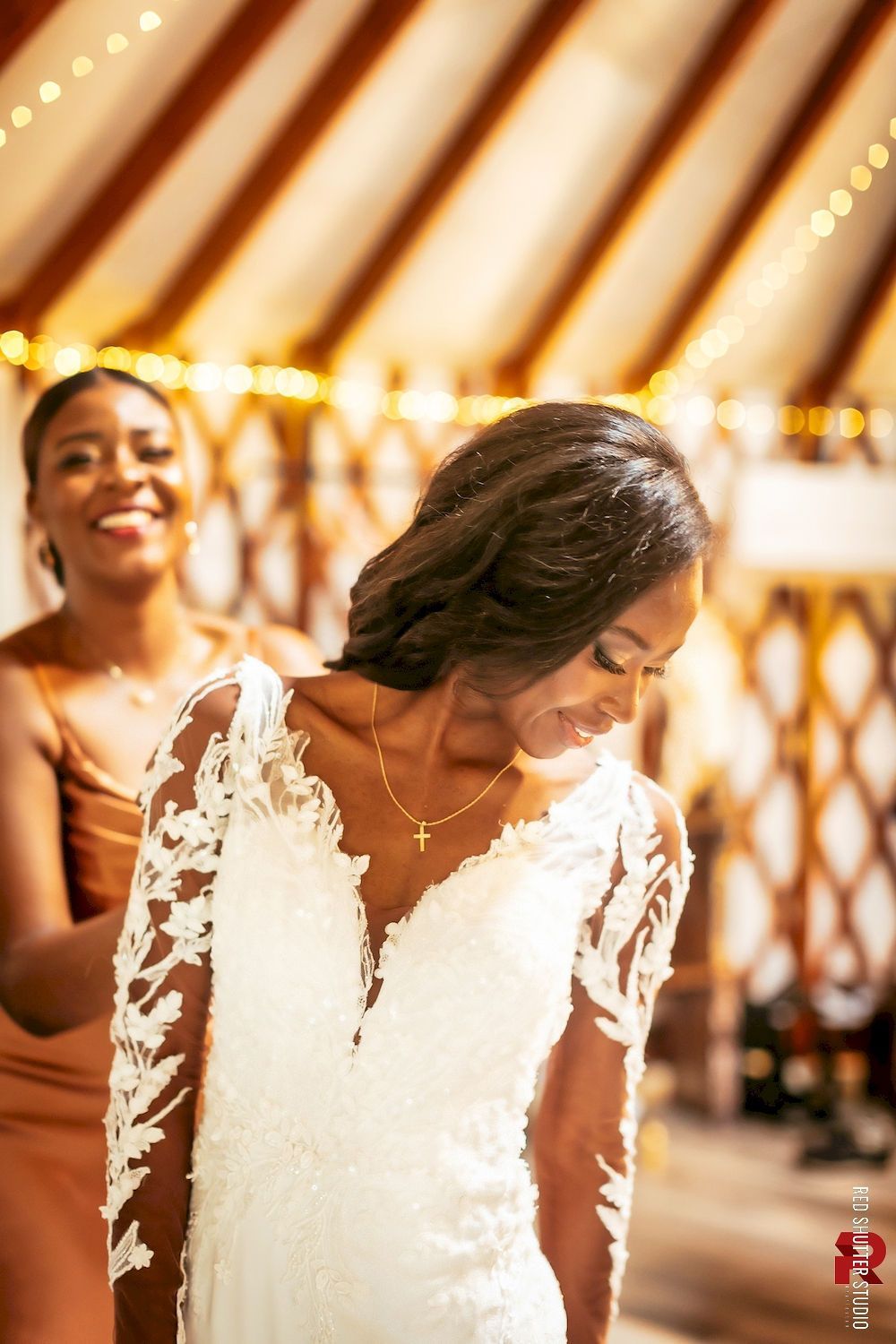 Bride in a lace white dress smiling with head bowed, while a guest smiles behind her at a warmly lit wedding venue