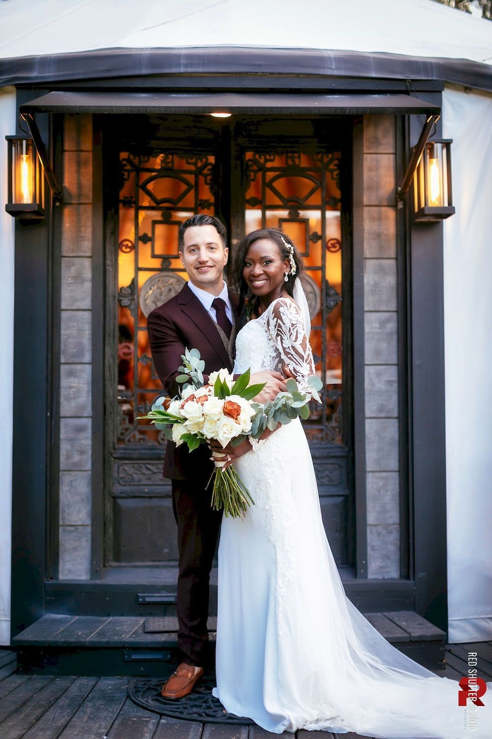 Wedding couple posing outside a doorway with lanterns, holding a bouquet, smiling in formal attire.