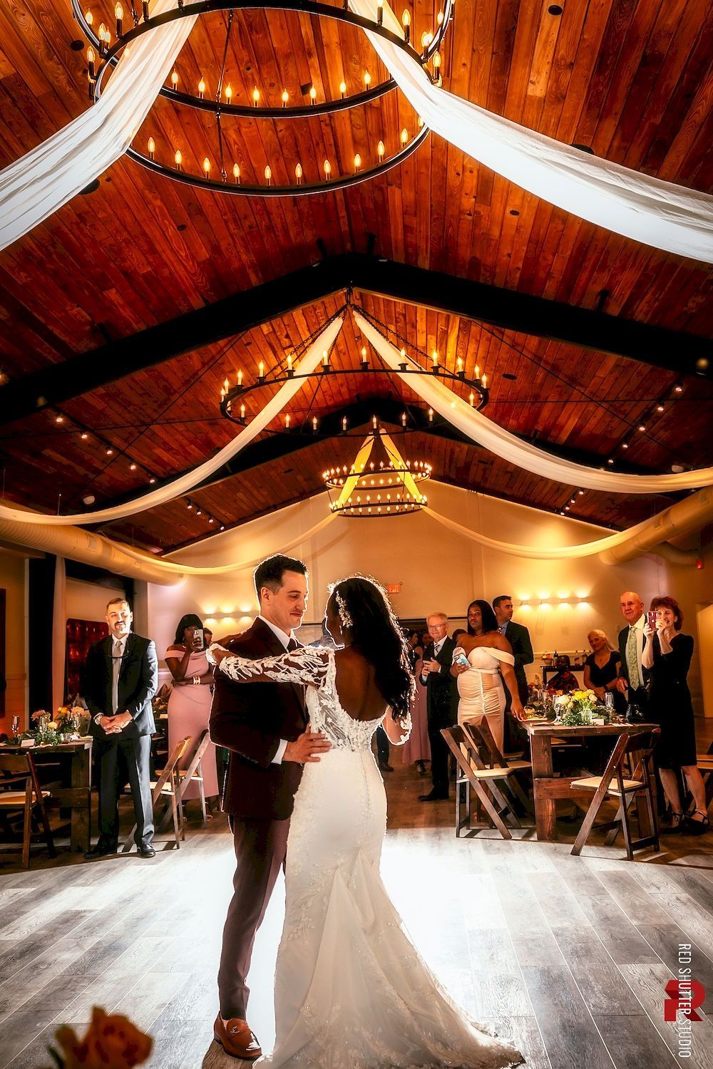 Couple dancing in a warmly lit rustic hall with draped ceiling and guests seated at tables.