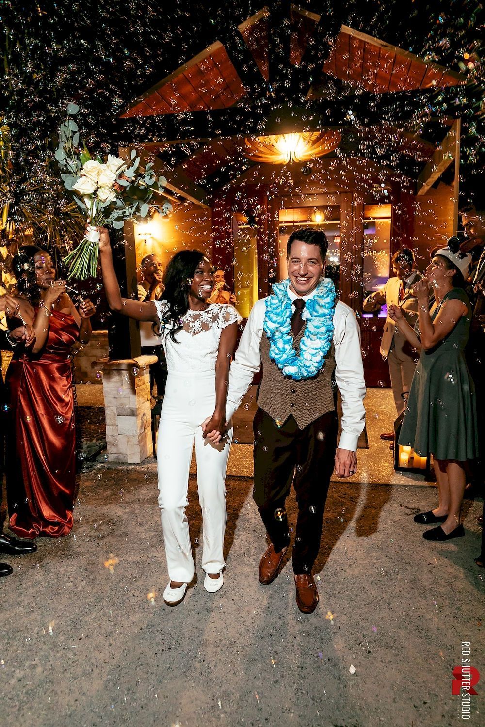Two people walk under confetti at a warmly lit celebration venue, surrounded by guests.