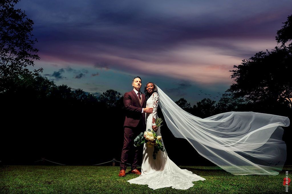 Couple in wedding attire standing on grass at dusk, bride’s veil flowing dramatically in the wind.