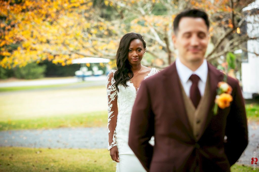 Bride in white stands behind groom in brown suit at an outdoor wedding with autumn trees.