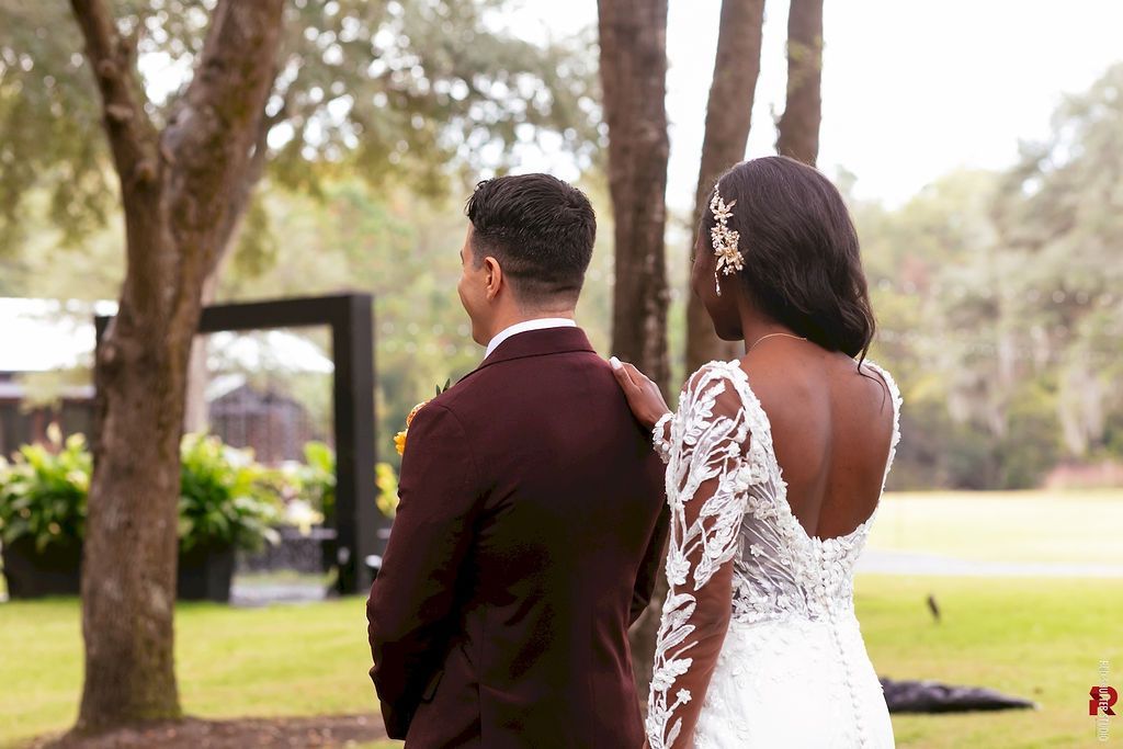 Couple in wedding attire standing outdoors beneath trees in a sunlit garden