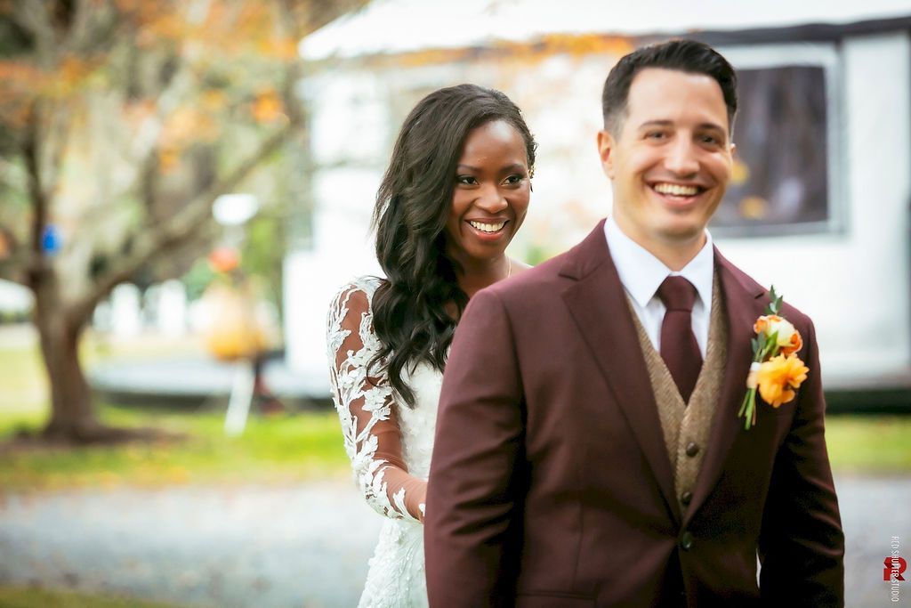 Newlyweds smiling outdoors, with bride in white lace dress and groom in burgundy suit and boutonniere