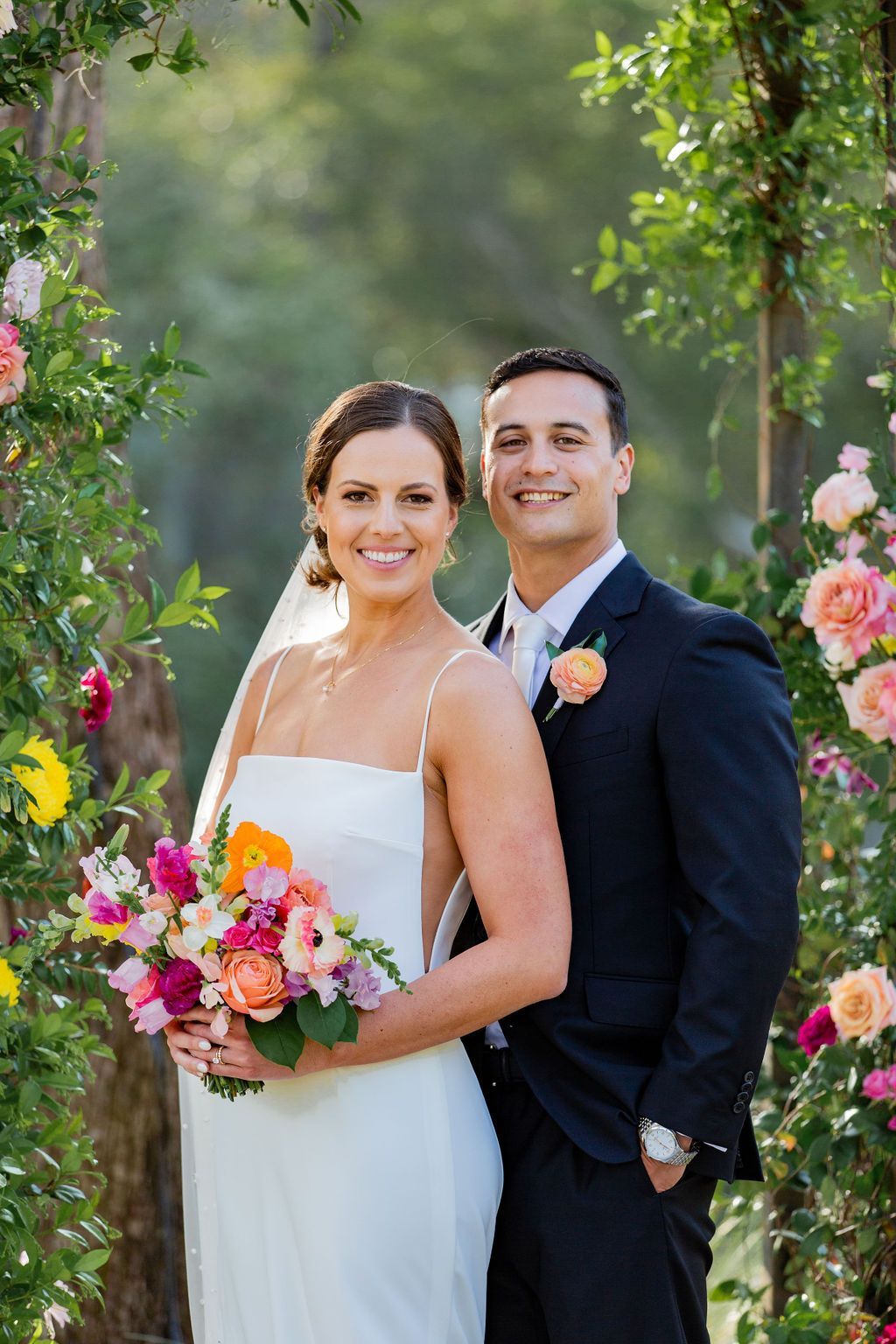 Bride and groom smiling in a floral garden, holding a colorful bouquet