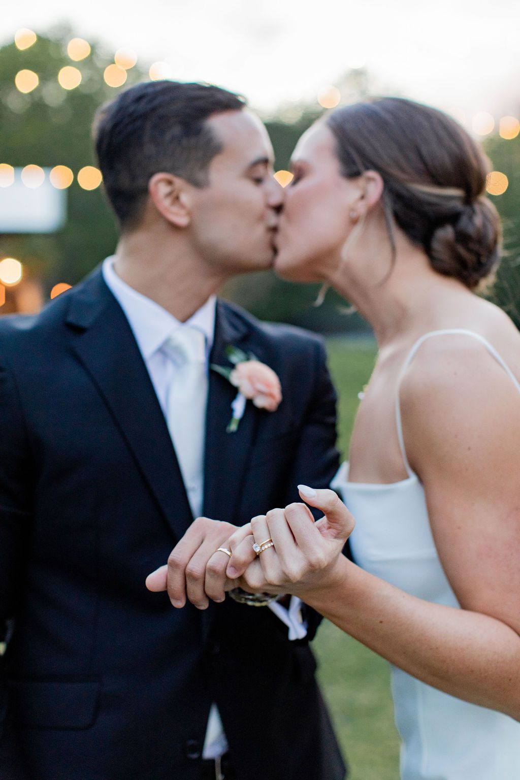 Two people in wedding attire kiss while holding hands outdoors at dusk.