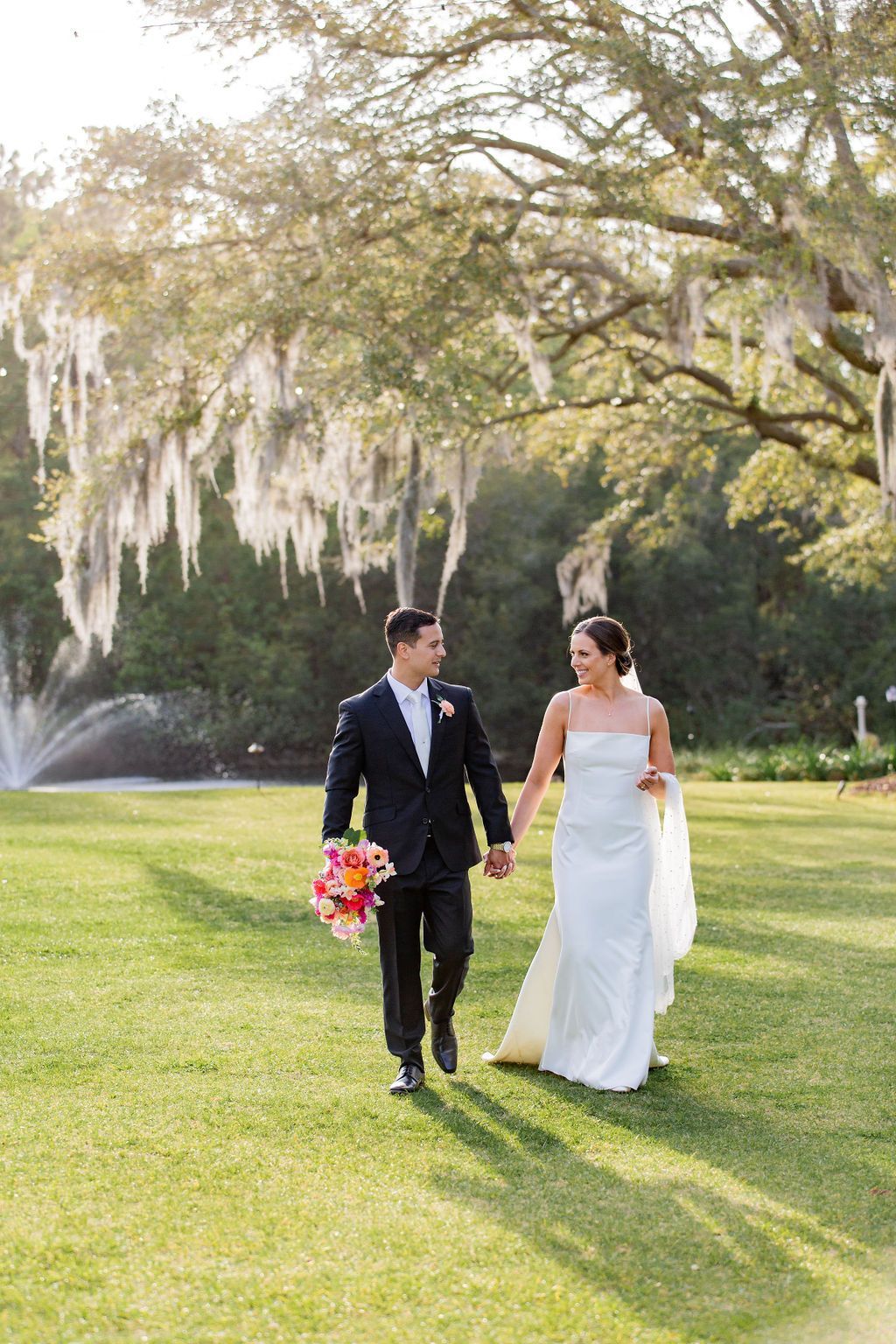Bride and groom walking hand in hand on a sunlit lawn with hanging moss trees behind them