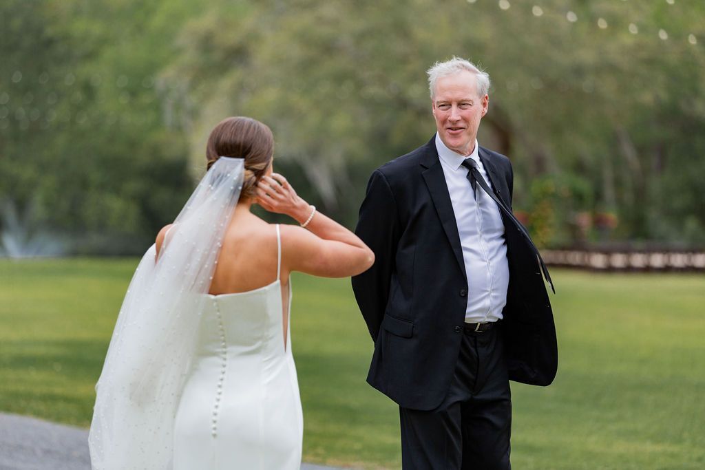 Bride in white dress and veil adjusts her hair while a man in a suit stands nearby on a grassy lawn.