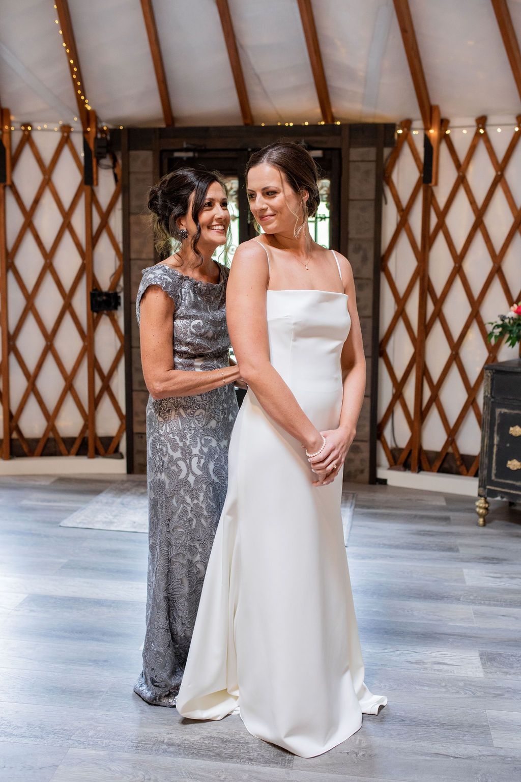 Two women in formal dresses posing indoors, one in white and one in silver, against a wooden lattice backdrop.