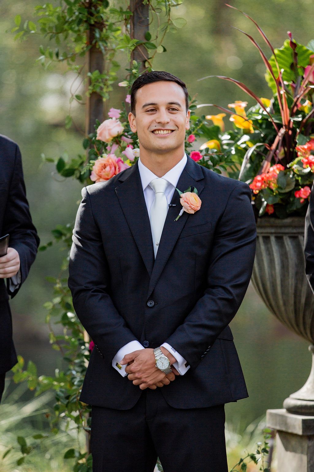 Man in a dark suit with a pink boutonniere standing outdoors beside floral arrangements
