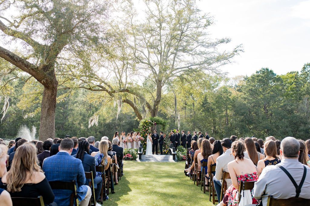 Outdoor wedding ceremony under trees, with guests seated on both sides of a grassy aisle.