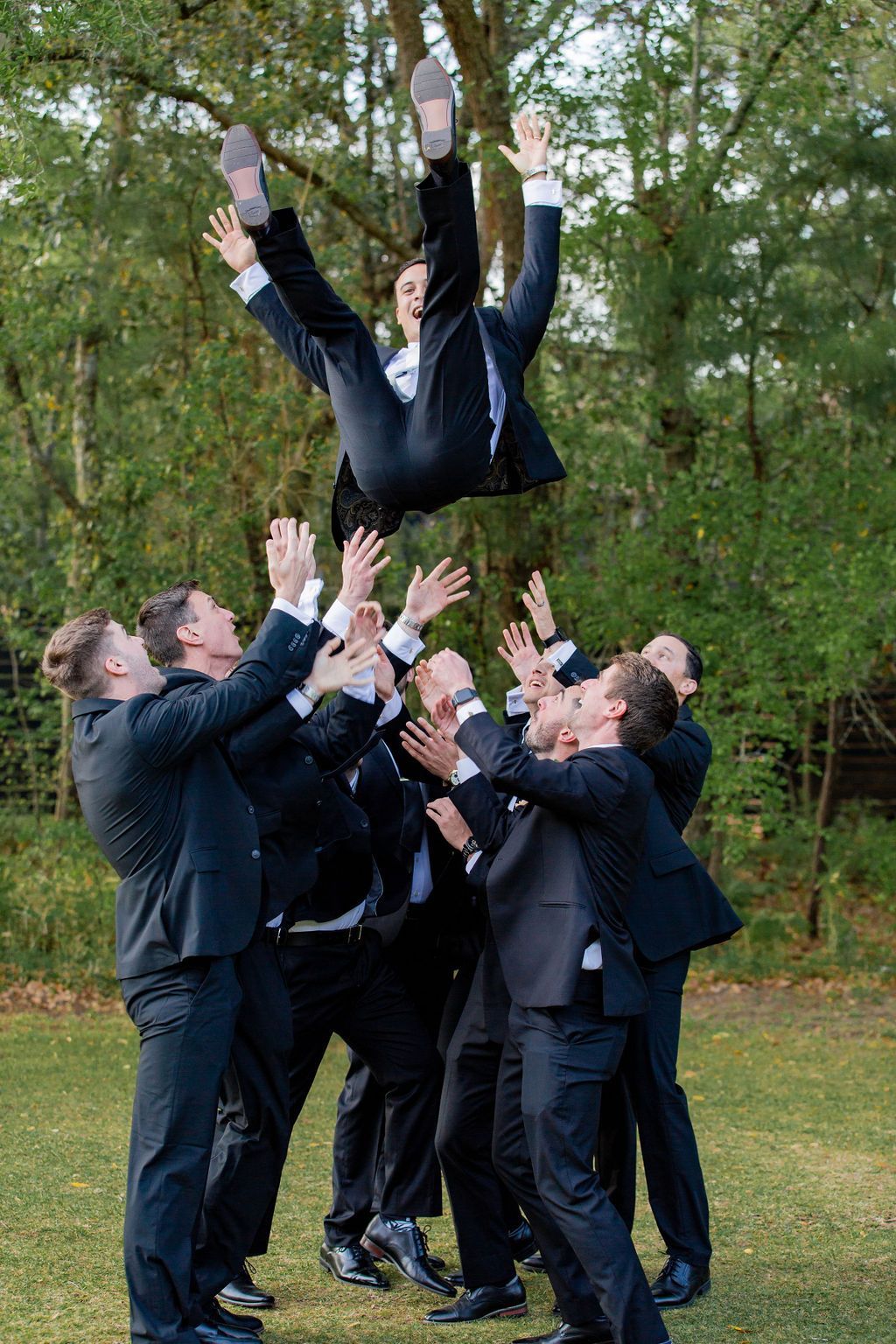 Men in dark suits tossing a colleague into the air in a wooded outdoor setting
