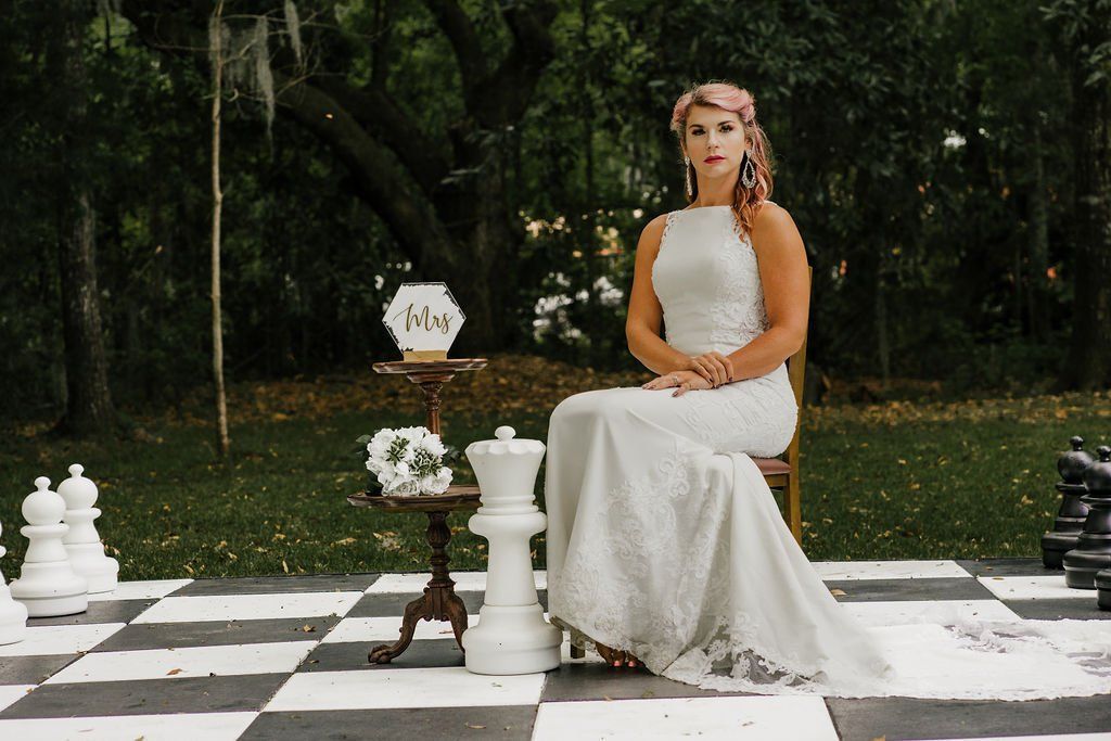 Woman in a white dress seated on a chessboard in a forest, with oversized chess pieces nearby.