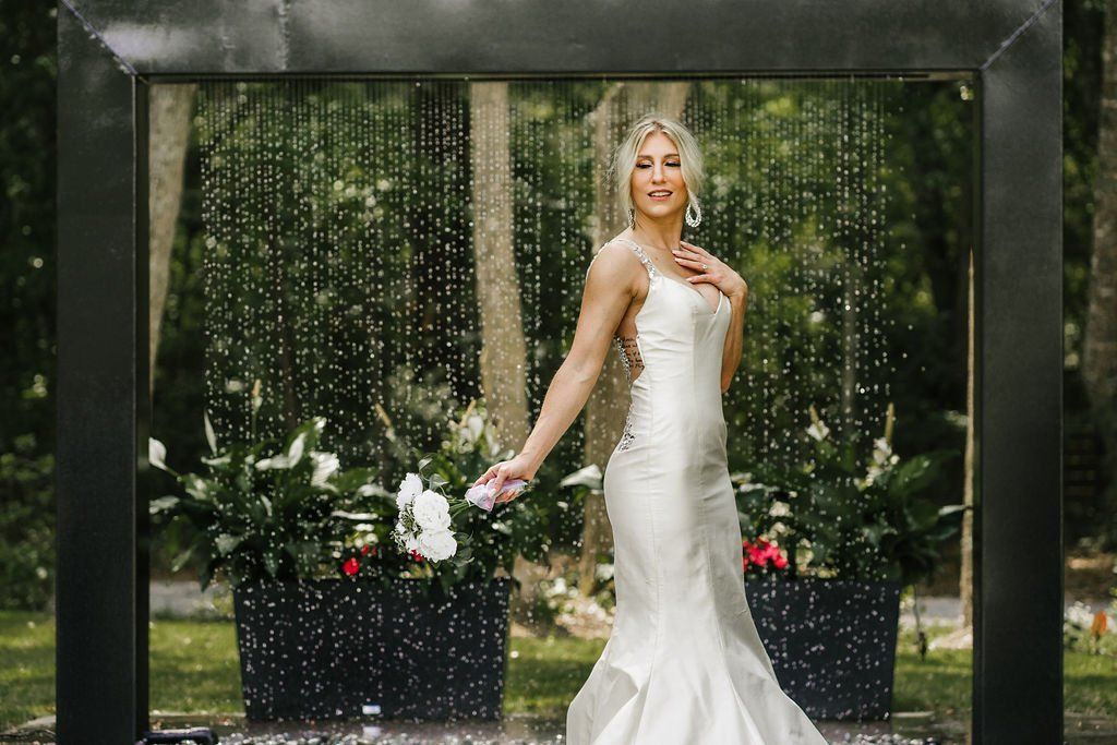 Bride in a white gown standing in a garden archway with flowers and greenery