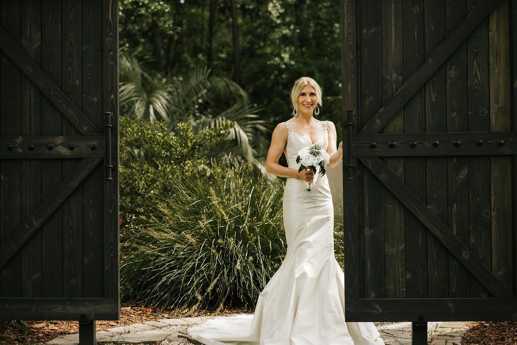 Bride in a white gown holding a bouquet, standing between large dark wooden gates with greenery behind.