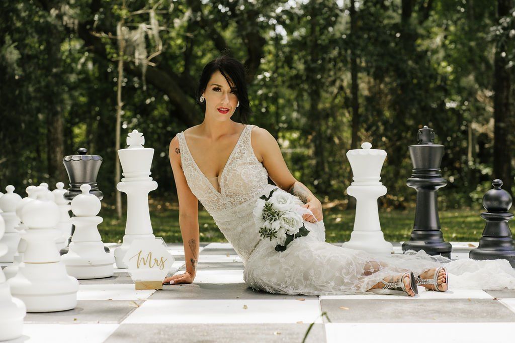 Bridal portrait of a woman in a white lace dress posing on a giant chessboard outdoors with white and black chess pieces