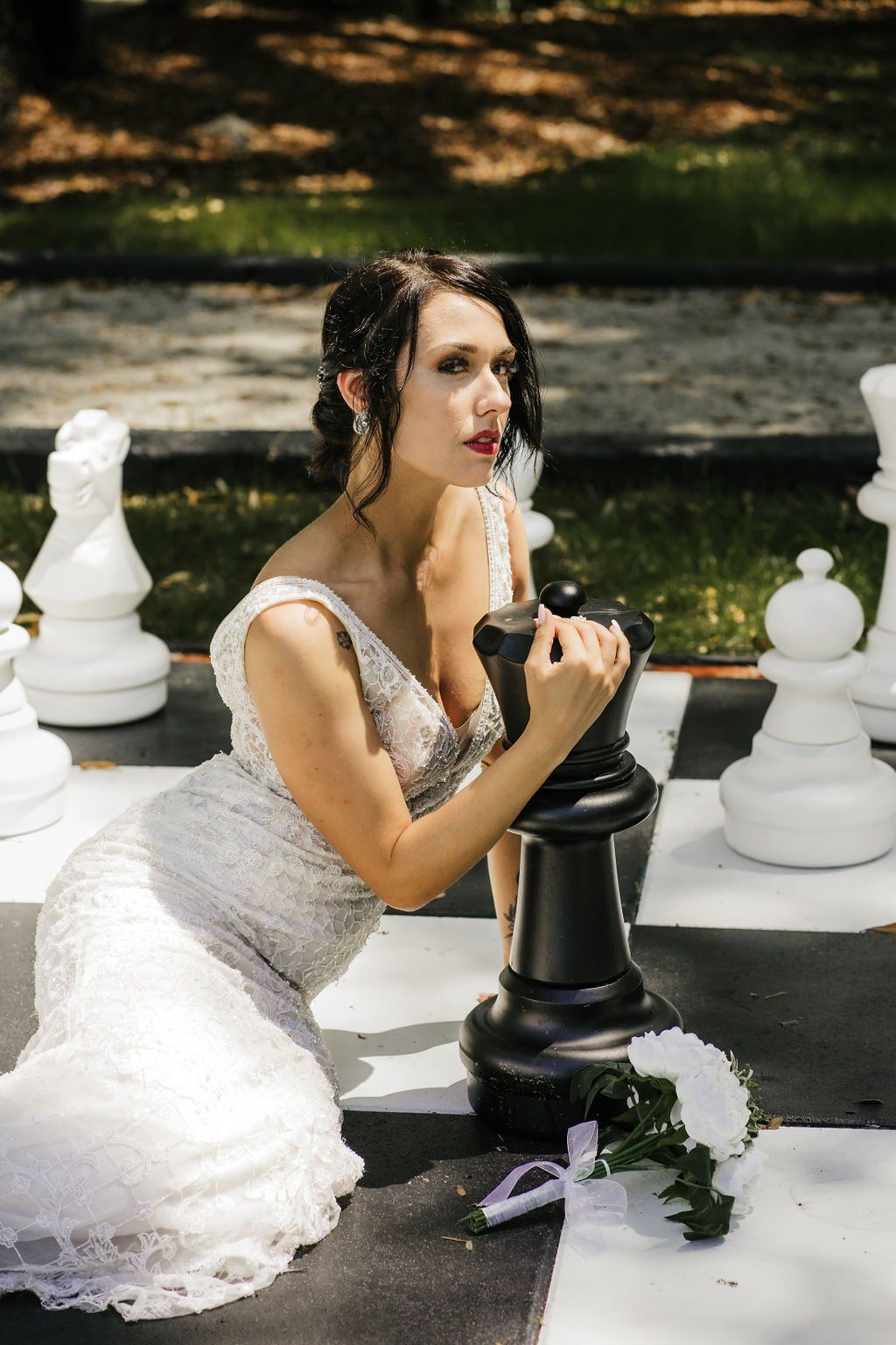 Bride in a white gown kneeling beside a black chess piece in an outdoor garden setting