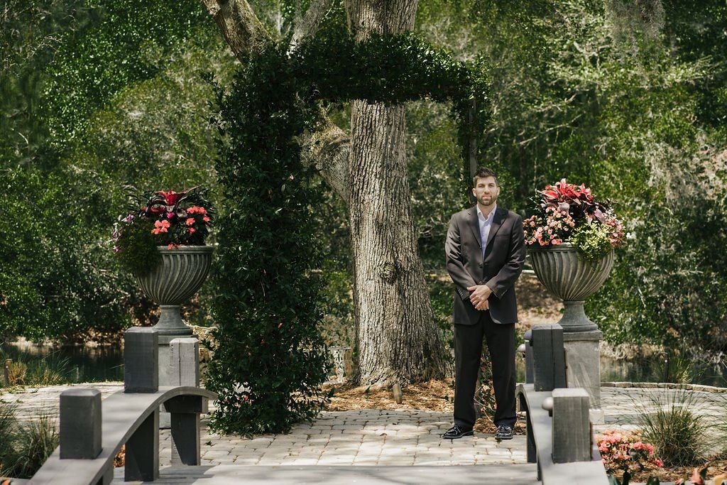 Man in a dark suit standing in a garden archway with red flower planters and greenery