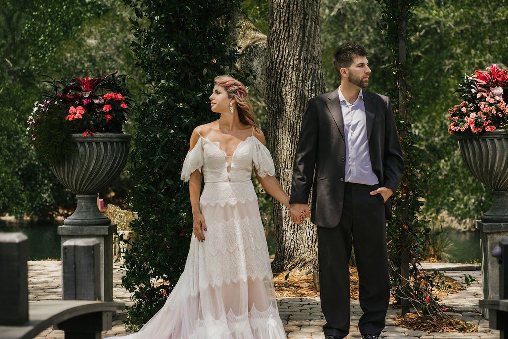 Bride in a white lace gown and man in a suit stand in a garden between flower planters.