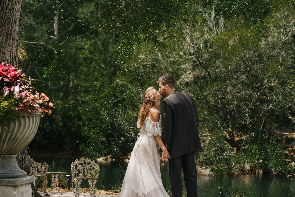 Bride and groom standing together by a pond in a lush garden, with flowers in the foreground