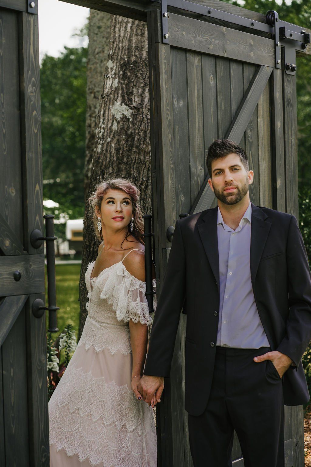 Bride and groom posing by a wooden gate outdoors, with trees and greenery in the background.