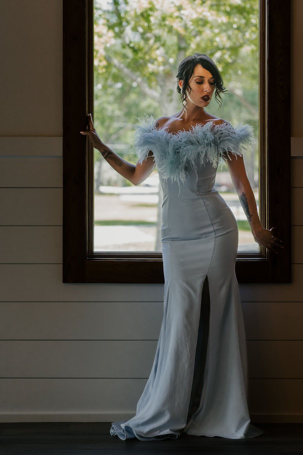 Woman in a white gown with feathered shoulders standing in a doorway with a bright window behind her.