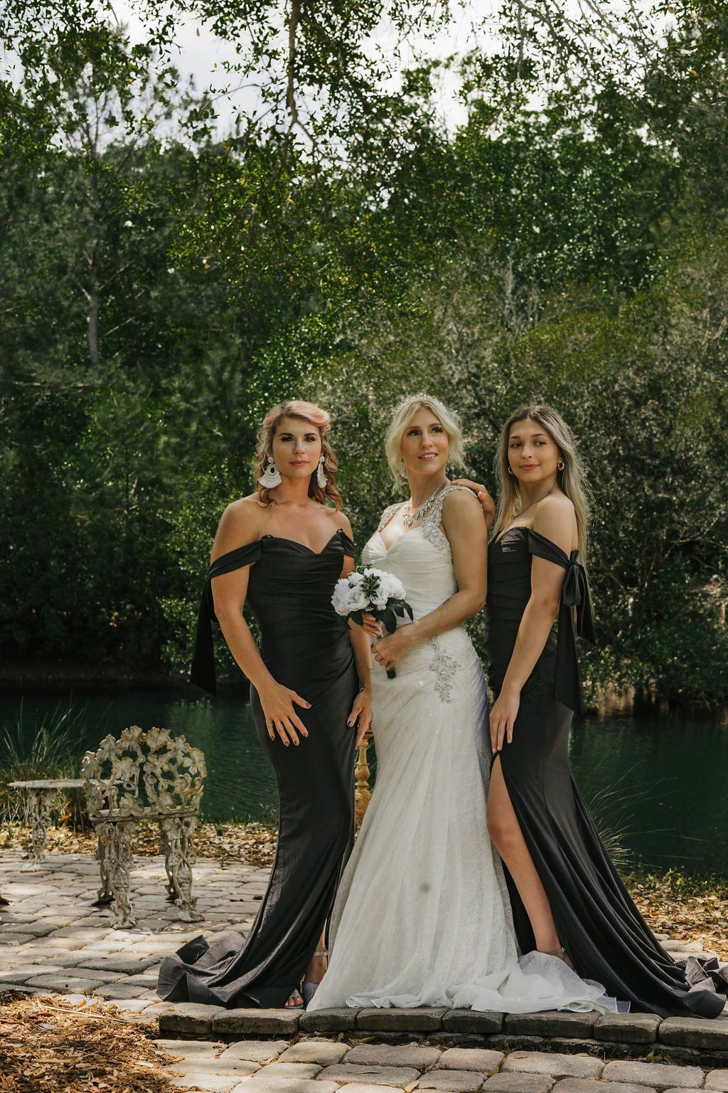 Three women in formal black and white dresses pose outdoors by water and greenery.
