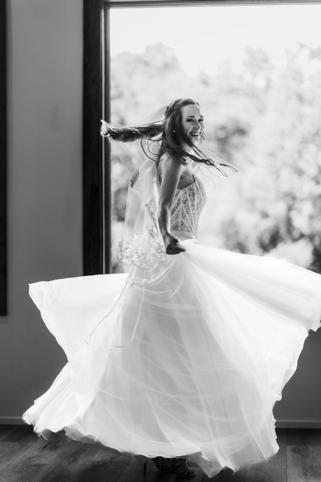 Bride in a flowing white gown twirls by a window, holding her skirt as it fans out gracefully.
