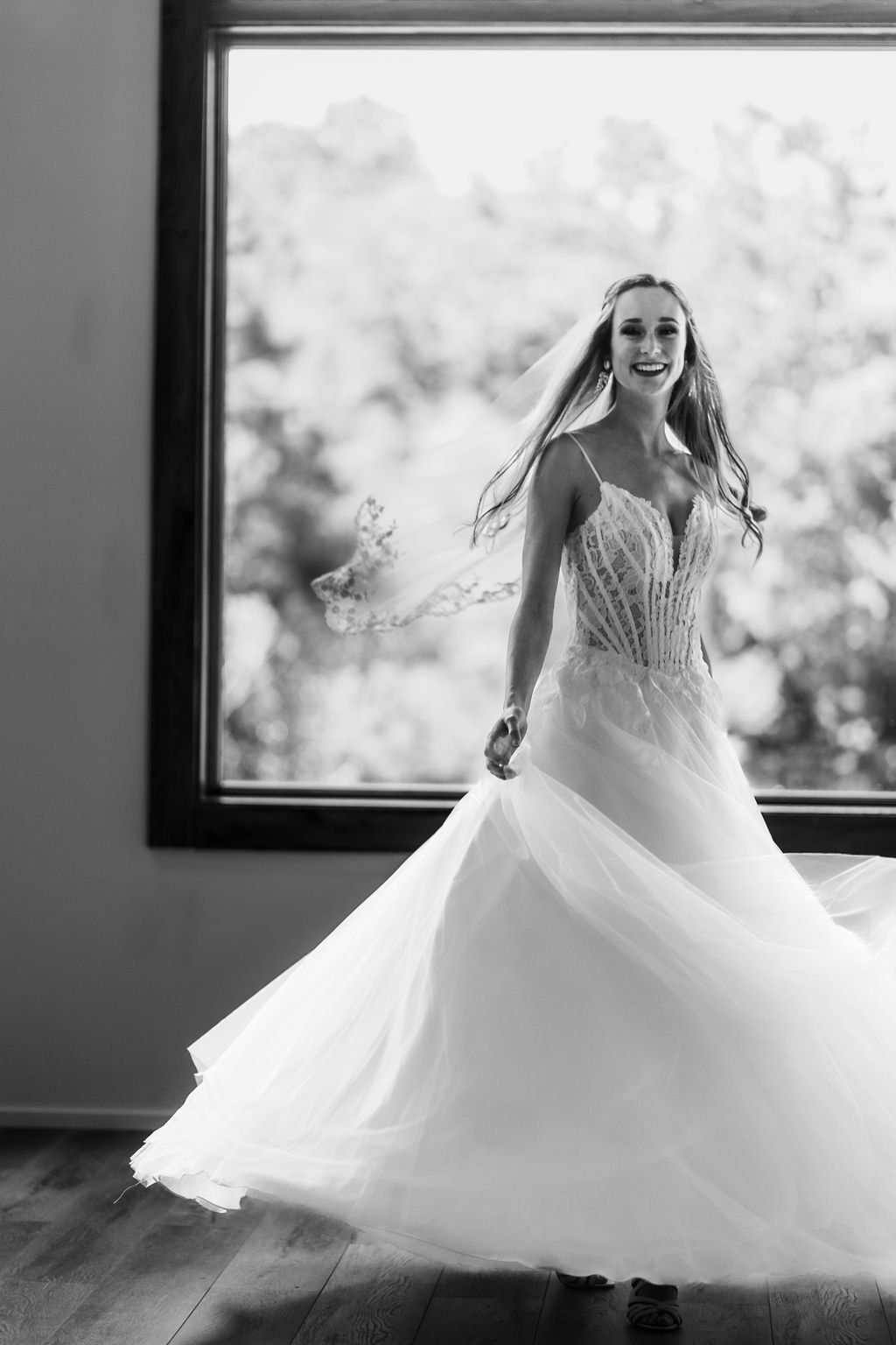 Bride in a white wedding gown standing by a large window, smiling in a bright room.