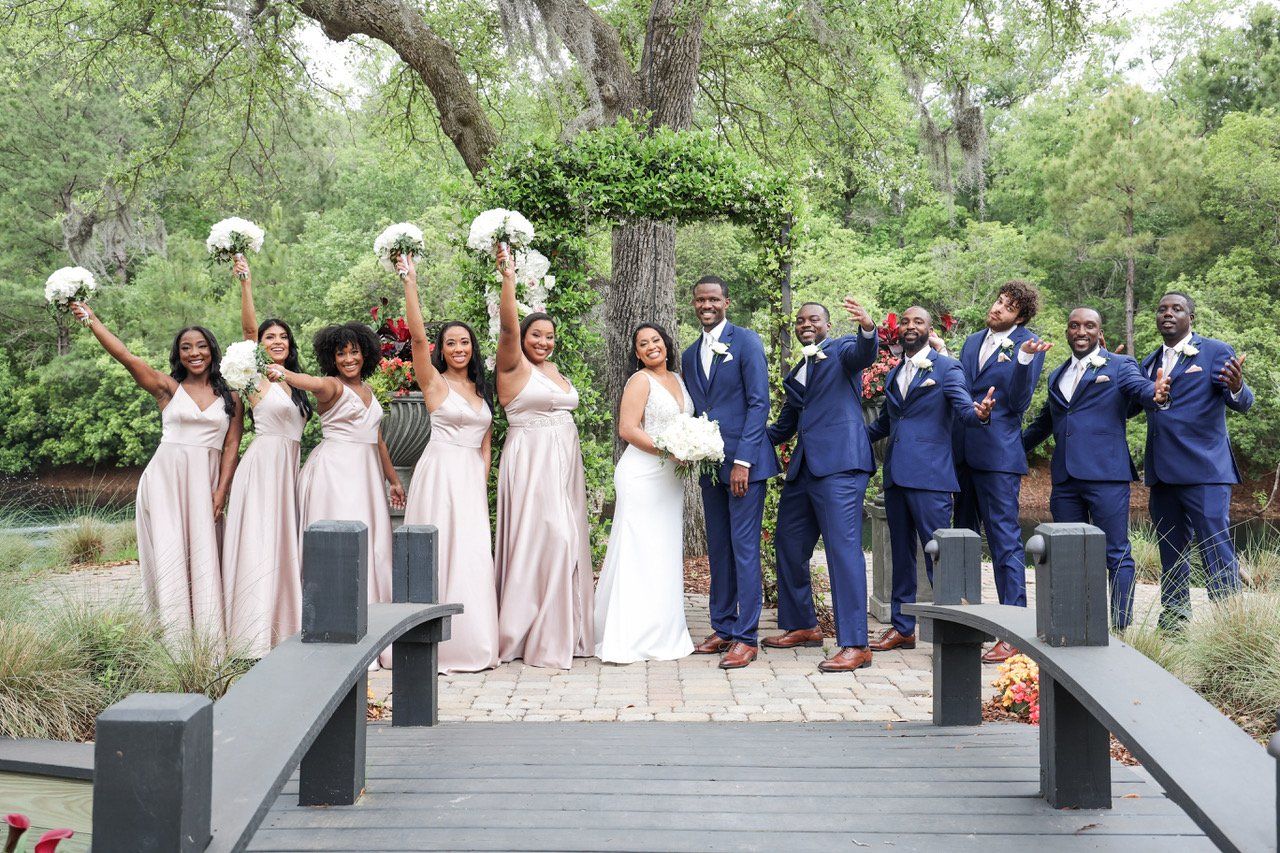 Wedding party on a bridge, bride and groom centered, bridesmaids in blush and groomsmen in navy raising bouquets.