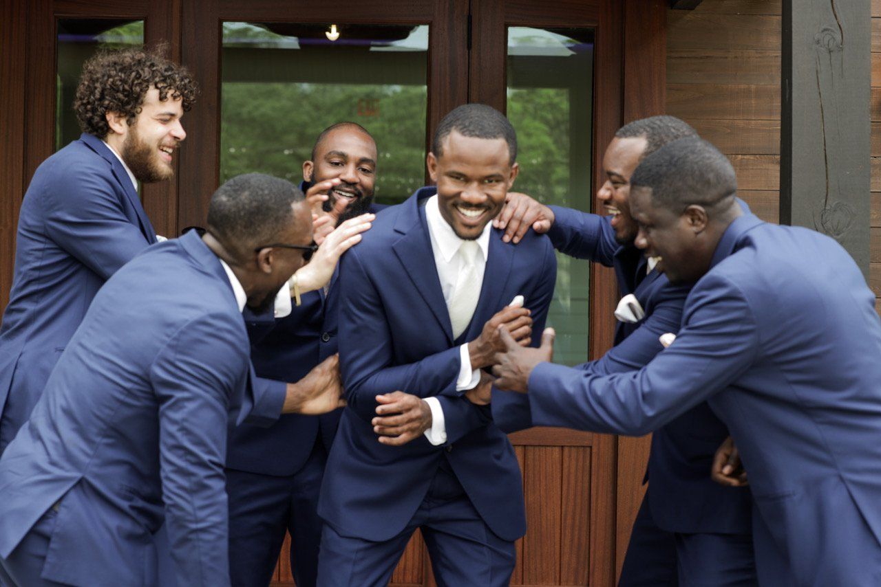 Five men in blue suits laughing and celebrating together outdoors by a wooden building