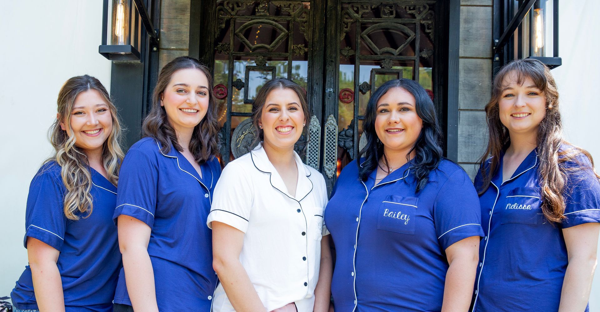 Five smiling team members stand together in front of a building entrance, wearing blue and white shirts.