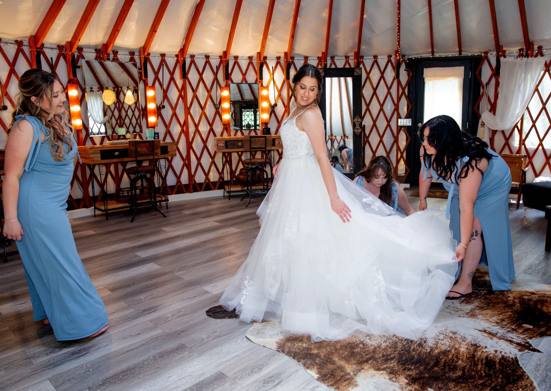 Bride in white gown with attendants in blue dresses inside a rustic yurt, adjusting the dress train