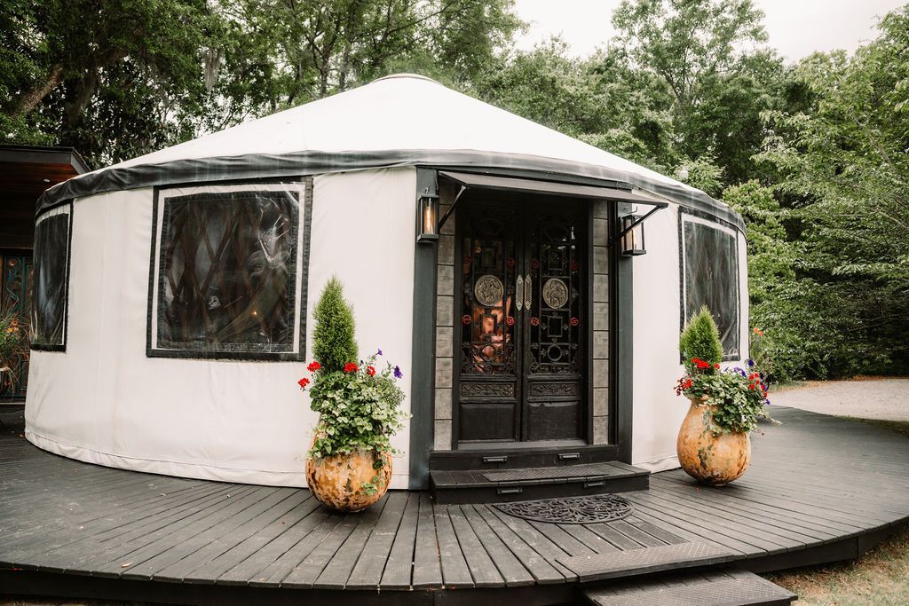 Round white tent with a black entrance, surrounded by flowers on a wooden deck in a wooded setting