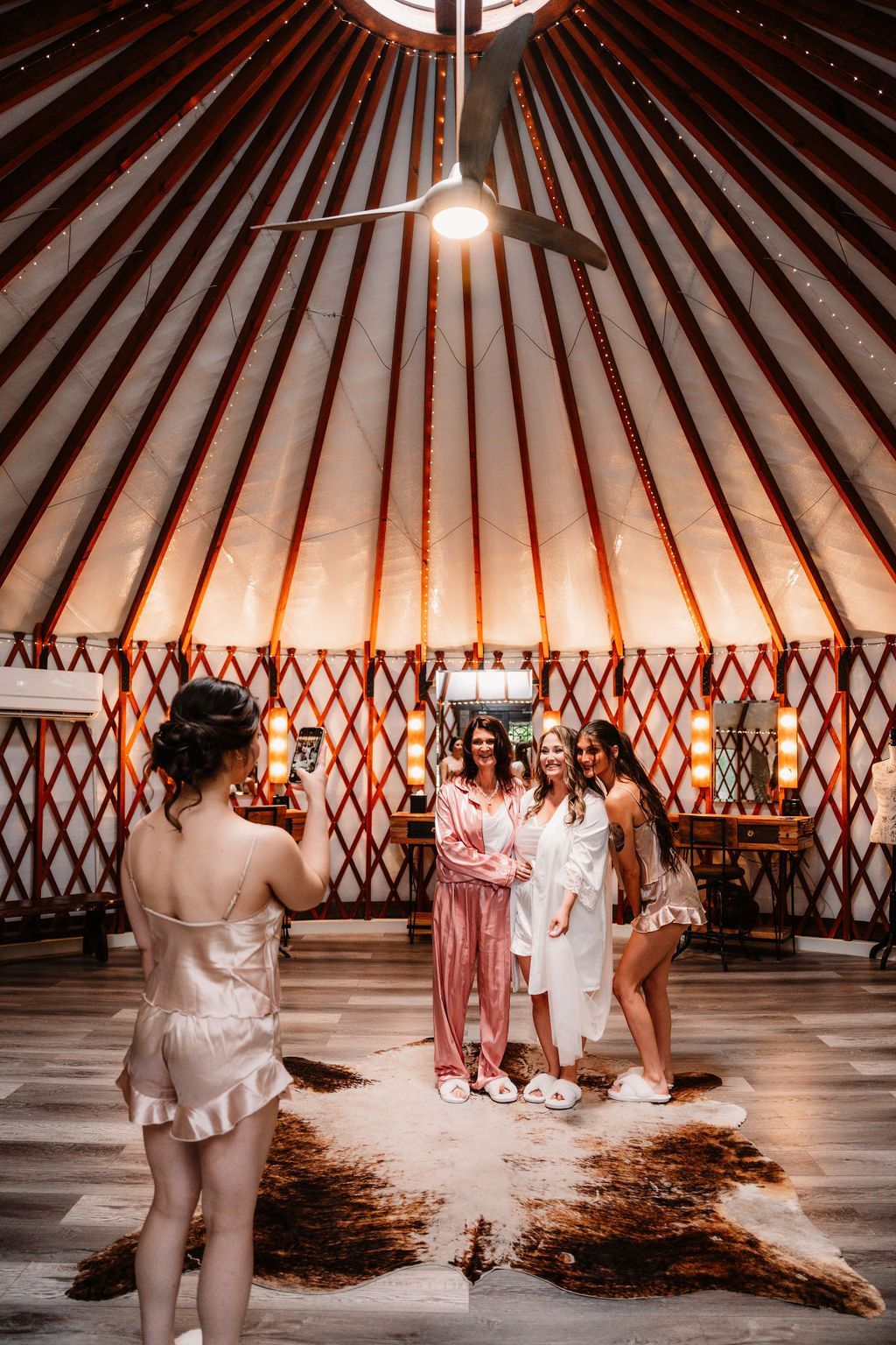 Three people in white stand inside a warmly lit yurt with patterned walls and fur rugs on the floor.