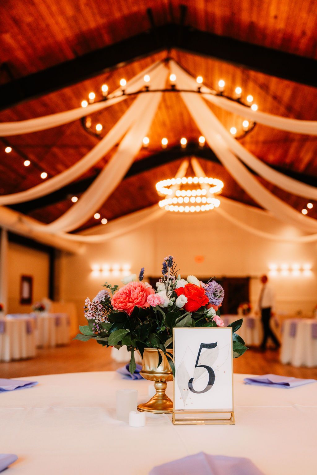 Wedding reception table centerpiece with flowers and table number 5 in a warmly lit hall