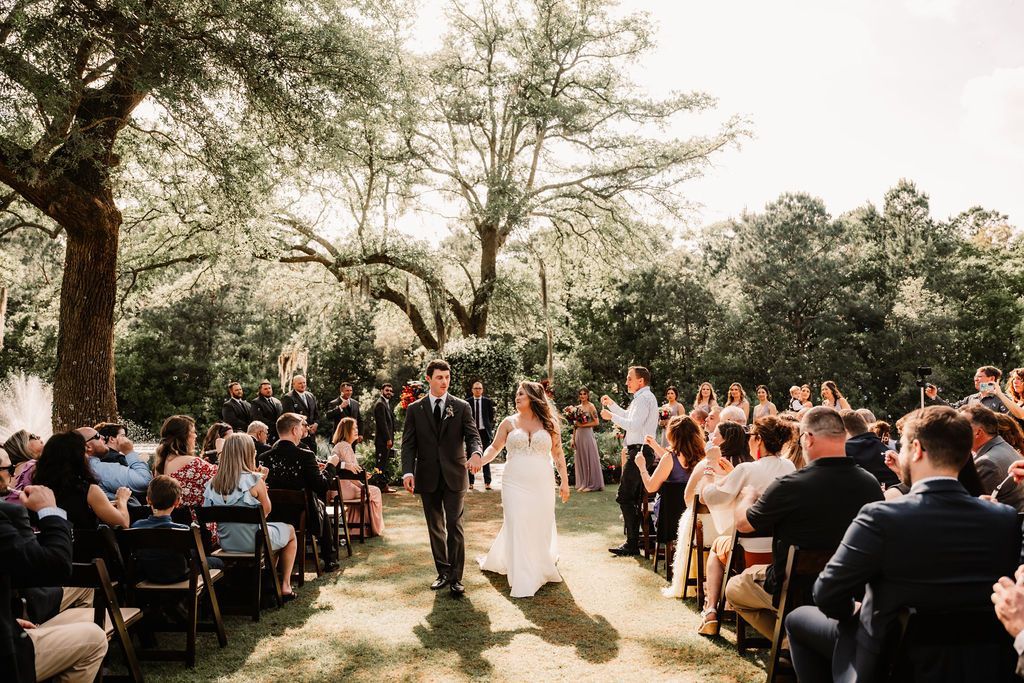 Outdoor wedding ceremony under trees with bride and groom walking down a lawn aisle past seated guests.