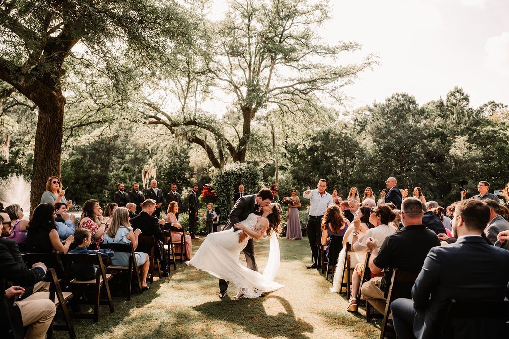 Outdoor wedding ceremony with bride and groom under trees, guests seated on both sides.