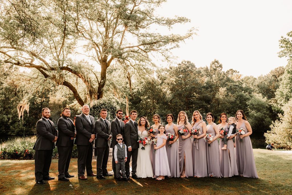 Wedding party posing outdoors under large trees, with bridesmaids in lavender dresses and groomsmen in dark suits.