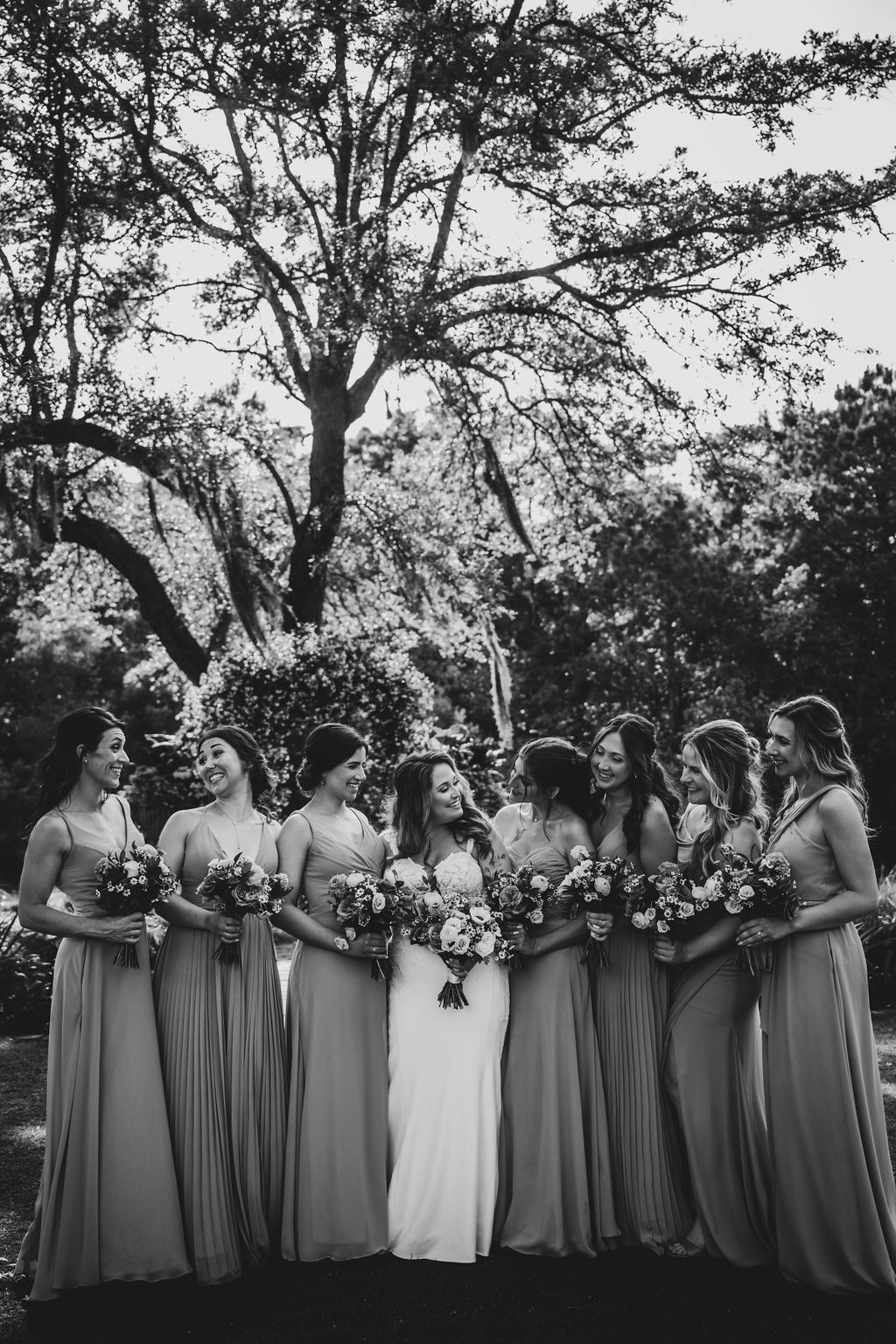 Bridal party in long dresses holding bouquets outdoors beneath a large tree