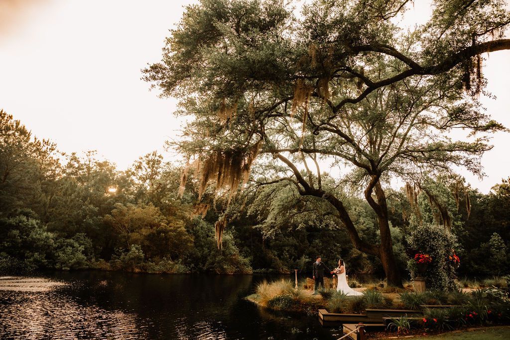 Riverside park with large trees, a calm pond, and two people sitting on the grass near the water